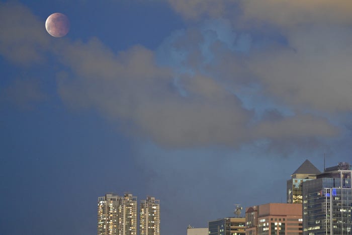 Half eclipsed moon in blue sky with low cloud cover, over a city skyline