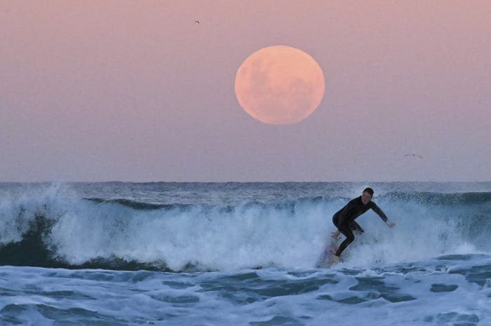 Close up of pink tinged moon at dusk near an ocean horizon with a surfer in the foreground