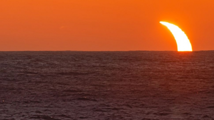 Partial crescent of white light peaks up from ocean horizon in red sky.