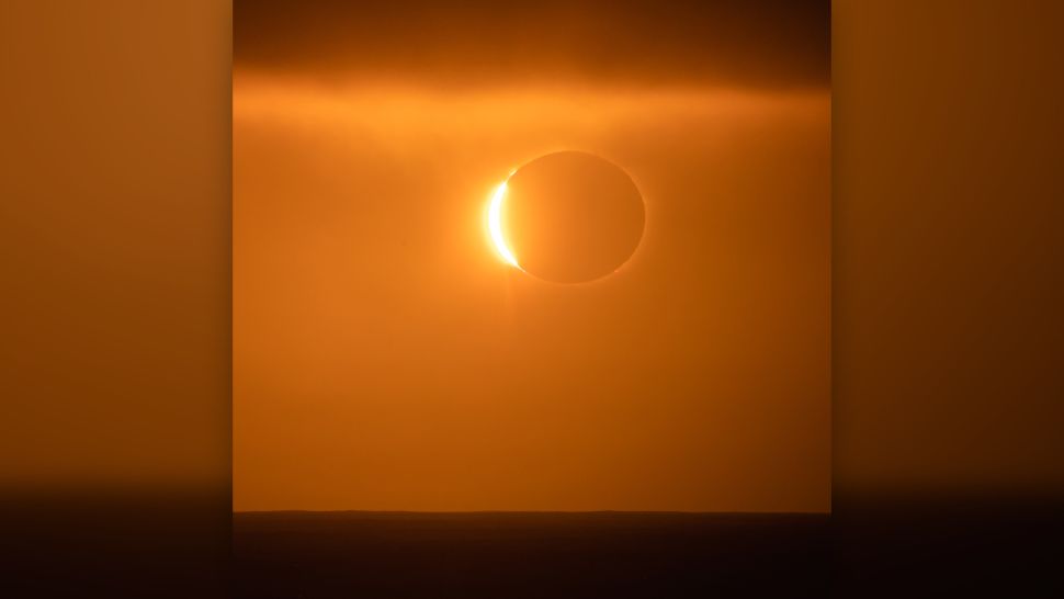 Sunlight oozes out from one side of sun blocking moon in red sky.