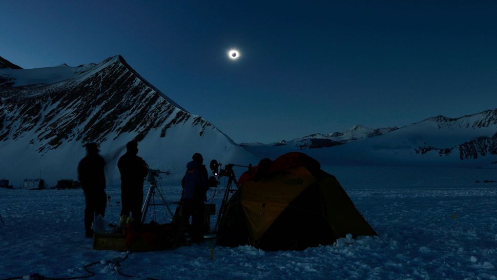 Three people with equipment and tent in front of mountain and eclipsing moon in dark blue sky.