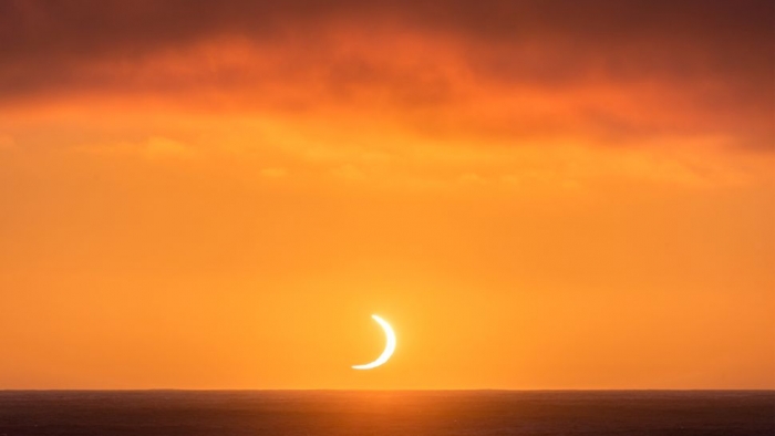 A Crescent Moon Of White Light Under Fiery Clouds In The Red Sky Above The Sea.
