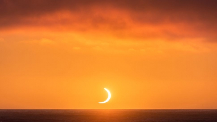 Crescent of white light, beneath fiery clouds in red sky over the sea.