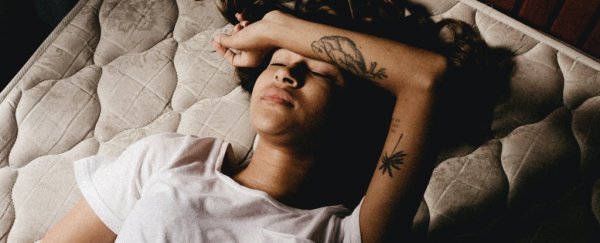 Woman with long dark hair and arm tattoos wearing white tshirt lying on bed with forearm on forehead.