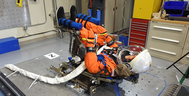 A mannequin in an orange spacesuit sitting in a chair.