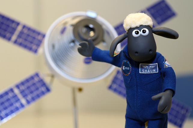 A sheep in a blue jumpsuit stands in front of a space capsule.