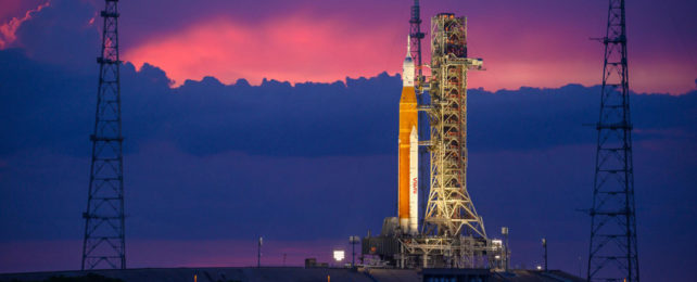 A rocket on a launch pad with purple and pink clouds in the sky.