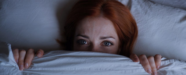 A woman in bed with her eyes open and her blanket pulled up past her nose.