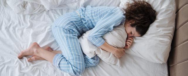 A woman in pajamas sleeping in the fetal position, curled around a pillow.