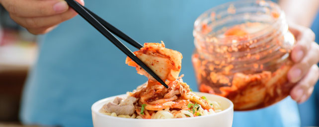 A person uses chopsticks to put kimchi on a bowl of noodles.