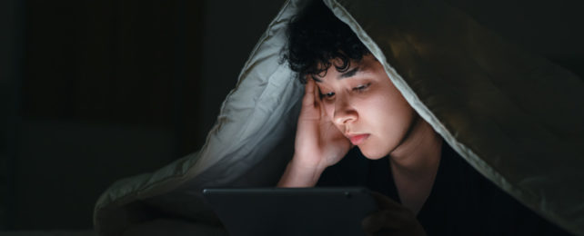 Teen stares at a tablet with a blanket pulled over their head.