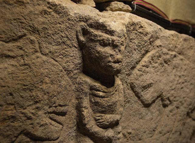 A close-up of an ancient carving of a man holding a phallus and two large cats approaching him.