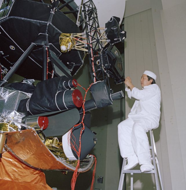 Engineer in white suit sitting on a ladder, working on Voyager 1 instrument.