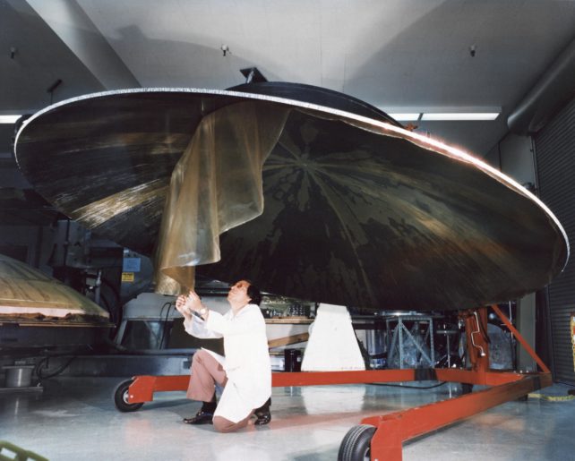 Engineer in white lab coat crouched beneath dish-shaped antenna of Voyager 1, peeling off a protective film.