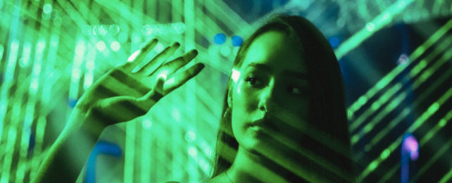 Girl standing amongst green beams of light, with hand raised.