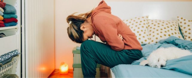 A woman sitting on a bed, doubled over in abdominal pain.