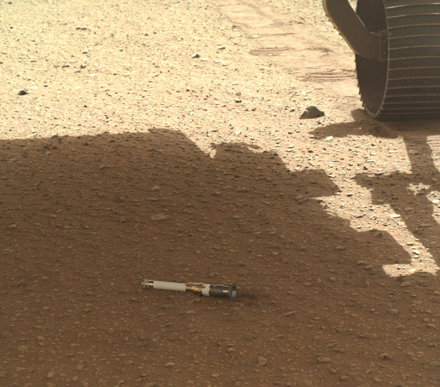 Un cilindro de almacenamiento de muestras en la superficie de Marte.  Visible en la esquina superior derecha está la Rueda de la Perseverancia.