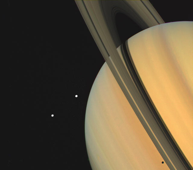 An image of Saturn showing its rings and two moons, which appear as bright white dots close to the planet.