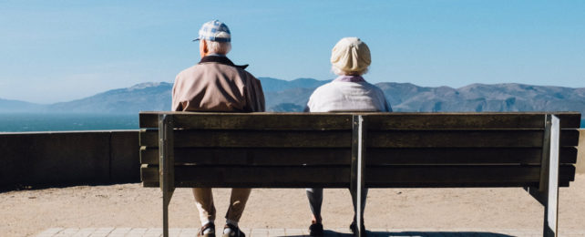 Two seniors sit on a bench looking out over the ocean.
