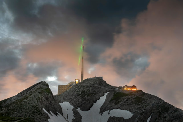 Green laser beaming up into cloudy sky above telecommunications tower on the summit of a snow-capped mountain.