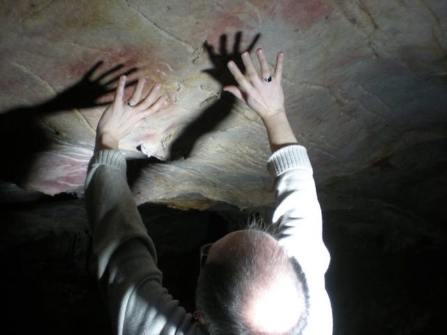 Man standing beneath cave roof with hands raised to highlight the position of hand stencils in illuminated cave.