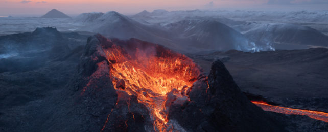 Volcano spewing red-hot lava in foreground of cloud-covered moutains.