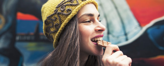 young woman with long brown hair in yellow beanie bites into a block of chocolate