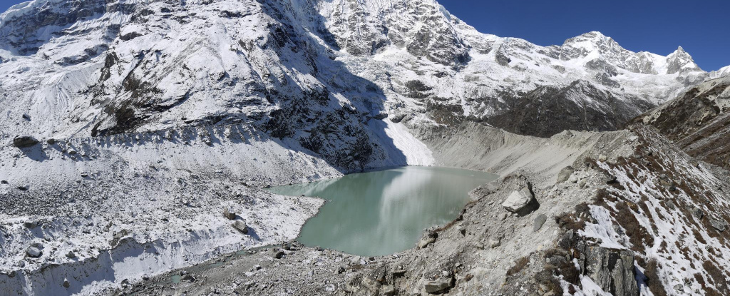 Cloudy blue-green glacial lake beneath snow-capped mountains on a blue-sky day.