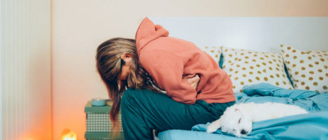 Girl in orange sweater hunched over in pain, sitting on the side of a bed with blue sheets.
