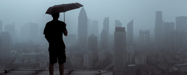 A man stands holding an umbrella while looking out at a misty skyline.