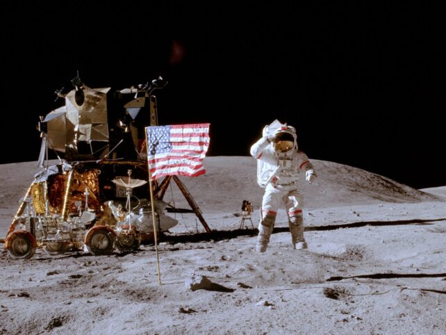 Seorang astronot memberi hormat pada bendera di tengah lompat saat berada di bulan mengenakan pakaian antariksa putih.