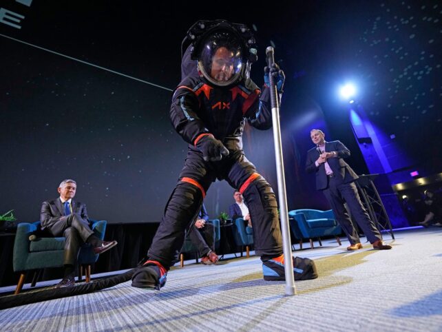 Ein Ingenieur hält in einem schwarzen Raumanzug-Prototyp ein Bein über dem Boden