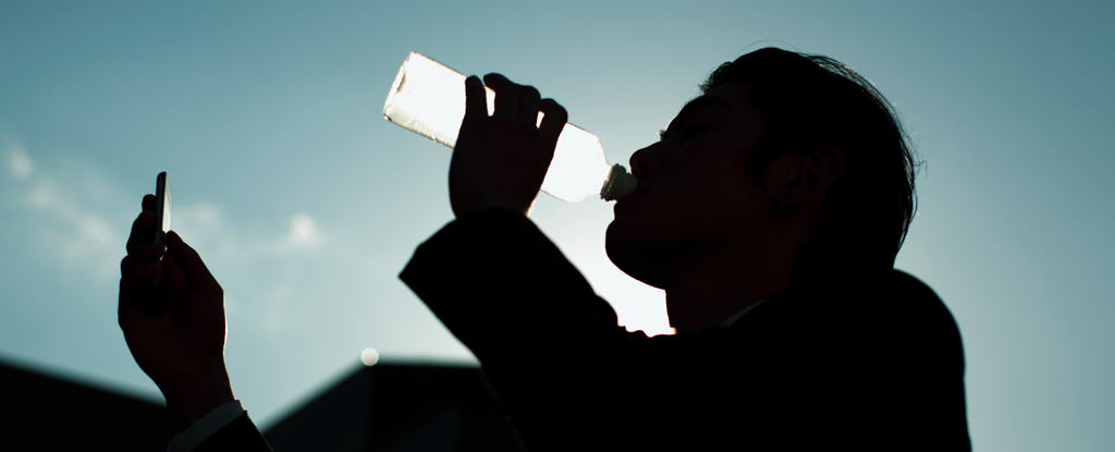 A man drinks water from a bottle while looking at his phone.