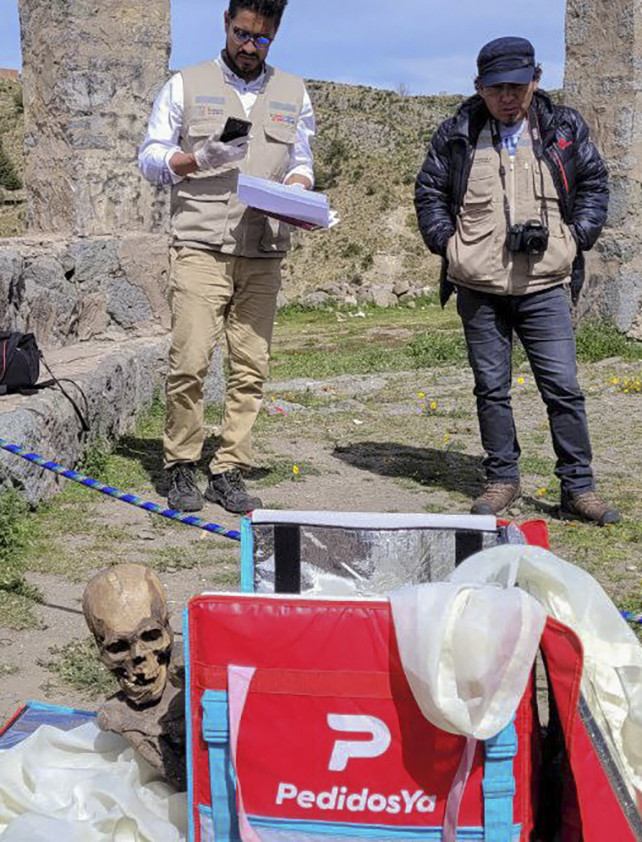 Two people look over the top of a red cooler bag, from which a mummy's skeletal head is emerging 