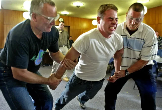 A police officer's painful reaction to being tasered during a training course on stun guns. A person is held on either side by two others, with a facial expression showing extreme discomfort. 