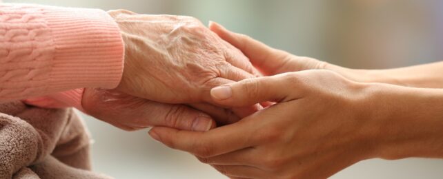 An older person's hands are held by younger hands in a close up image showing pink sleeves and a brown blanket in the corner.