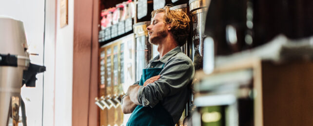 A sleepy barista leans against a shelf of coffee beans.