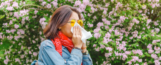 A woman blows her nose while walking past blooming plants.