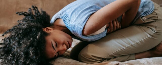 A women doubled over clutching her stomach in pain, wearing a blue top and tan pants.