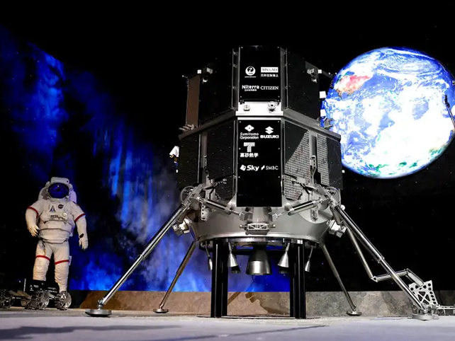 Model of the Hakuto on a stage next to a space suit with earth in the background