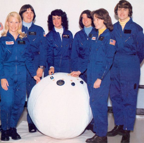 Six women astronauts in blue space suits standing around white rescue ball.