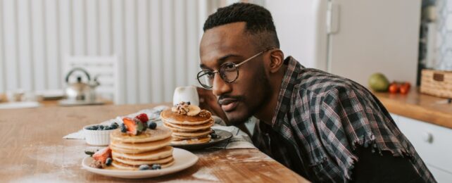 A man sitting at a table in a kitchen looking at two plates of pancakes.