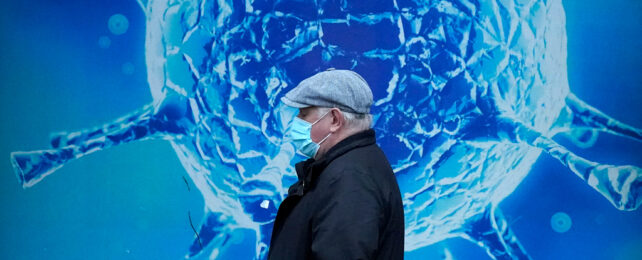 A man wears a surgical mask while walking past an illustration of a COVID-19 virus.