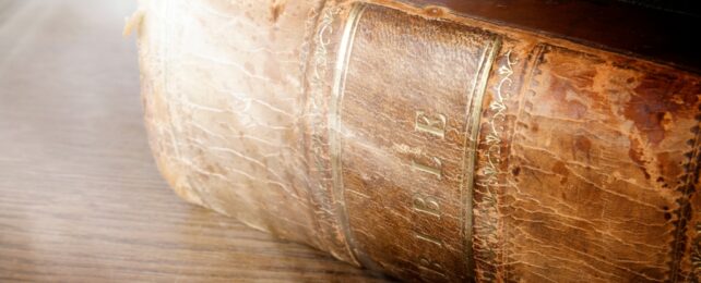 A close up of the spine of an old brown leather look worn bible with the title written in gold. Sun rays in background.