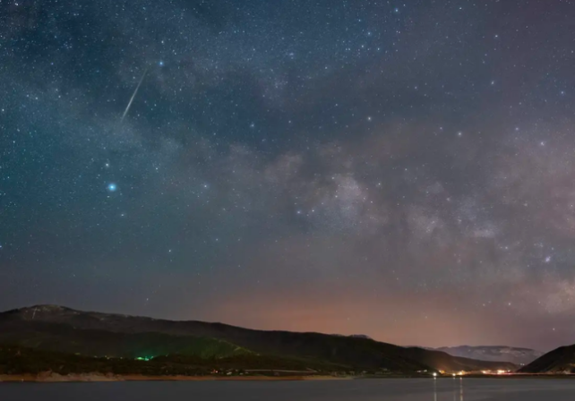 A dark starry sky over water in the foreground and land in the background. It looks like a 'shooting star' is visible