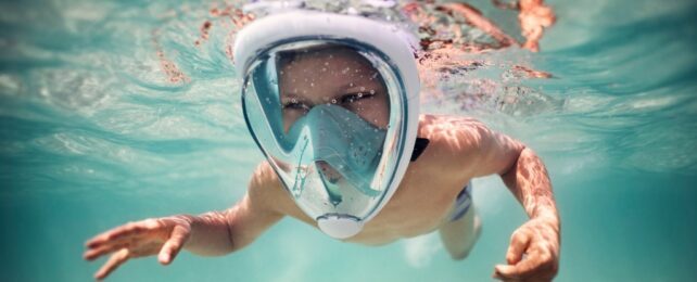 A boy swimming underwater towards the camera, wearing a full face snorkel mask.
