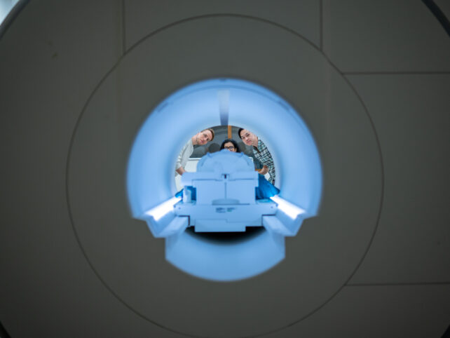 Three human faces peering through a fMRI scanner, that appears as a blue tunnel inside a grey tunnel.