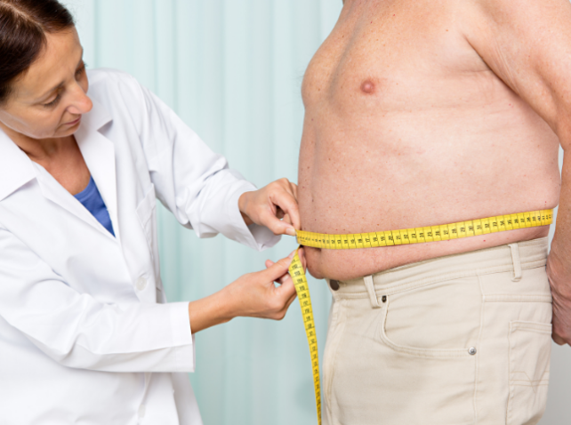 A woman in a white coat is holding a measuring tape around the stomach of a shirtless man wearing tan shorts. His face is not visible 