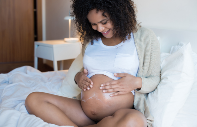 A person with dark curly hair sitting on a bed holding a pregnant belly