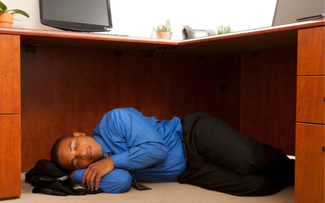 A man wearing office clothes sleeping under a desk during the day